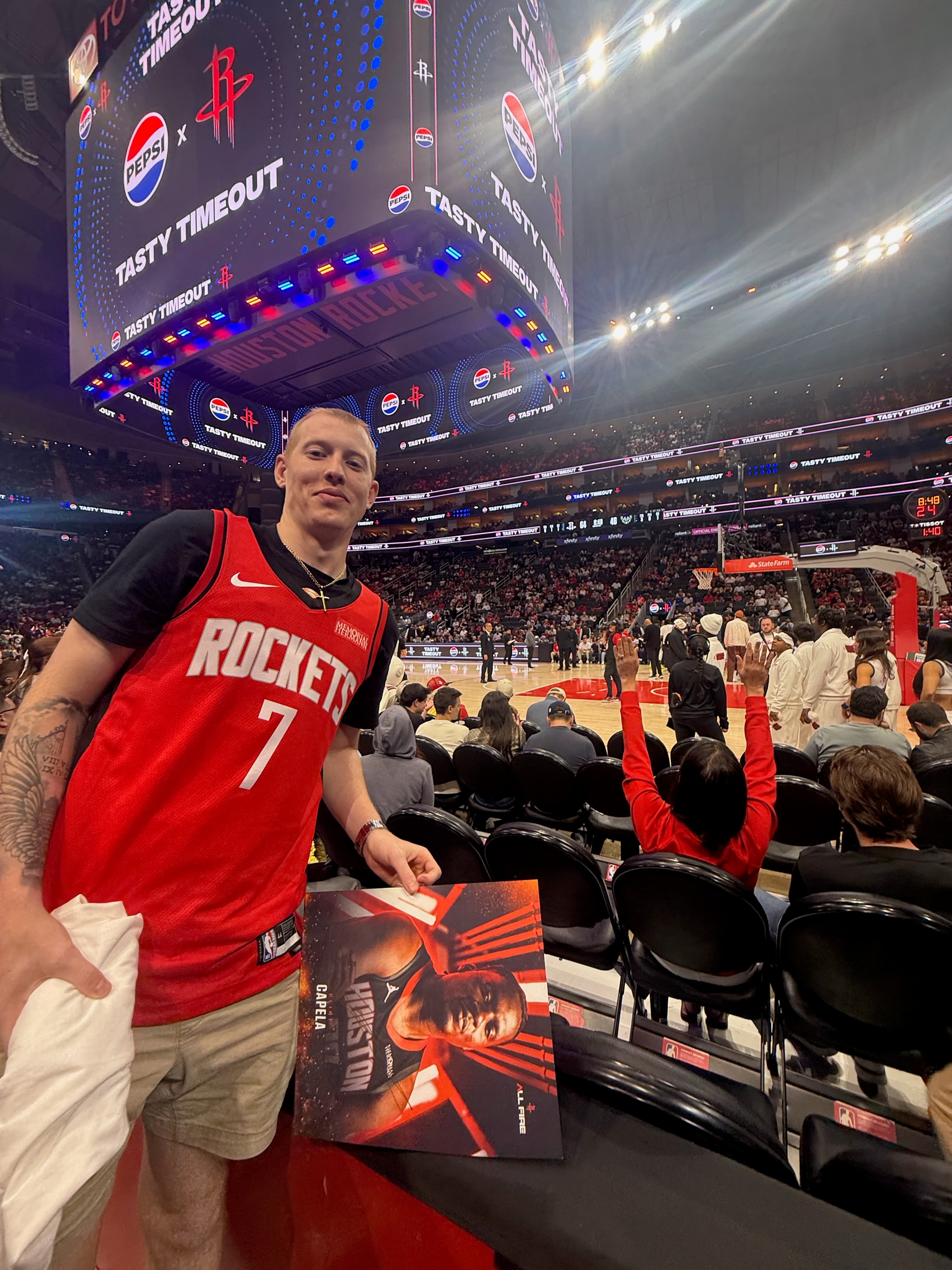 Fan in red Rockets jersey holds a Clint Capela poster inside a basketball arena.